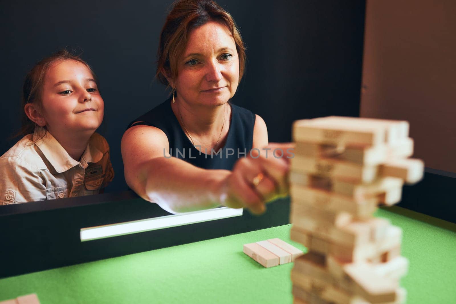 Mother playing jenga game with her daughter in play room. Woman removing block from stack. Unstable tower of wooden blocks. Game of skill and fun. Family time