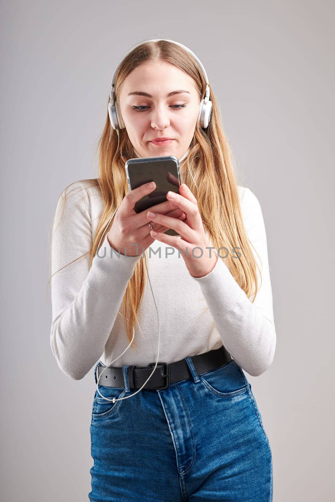 Young woman girl listening to music streaming content having fun watching video enjoying video chat talking with friends making gestures faces using smartphone earphones headphones standing over plain grey background