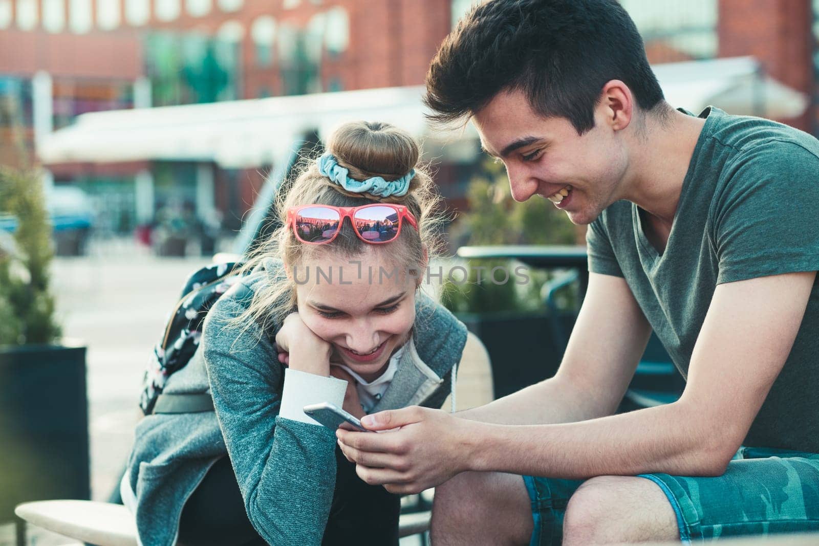 Couple of friends, teenage girl and boy,  having fun with smartphones, sitting in center of town, spending time together