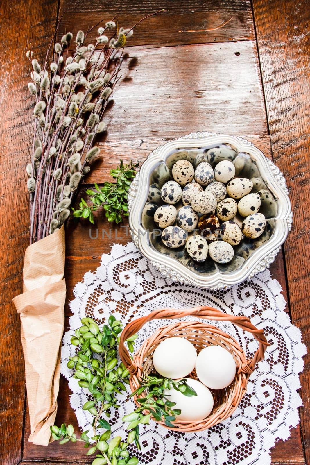 Easter composition of willow twigs with catkins, boxwood, quail eggs in a metal dish and easter basket with eggs, set on old wooden table. Catkins are wrapped up in paper. There is lacy napkin under the basket. Inside top shot. Vertical image.