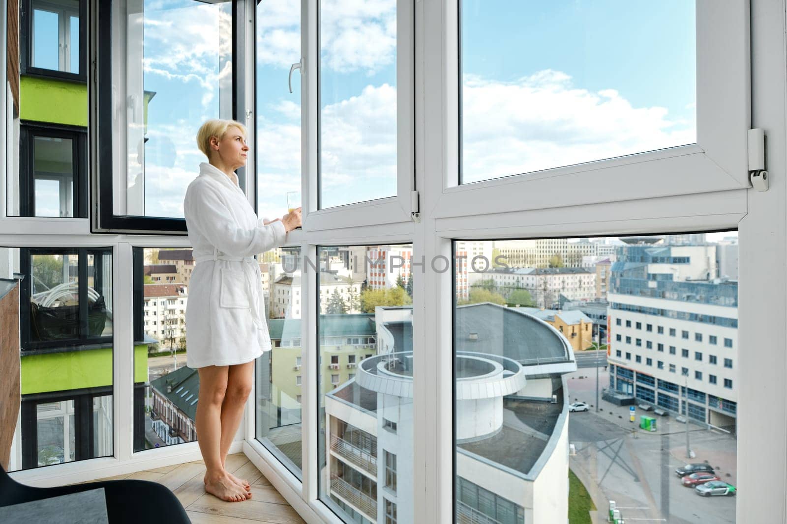 A mature, carefree woman stands barefoot in a bathrobe on her balcony enjoying an orange juice. Alone in her new high-rise apartment and overlooking the sunny city skyline.
