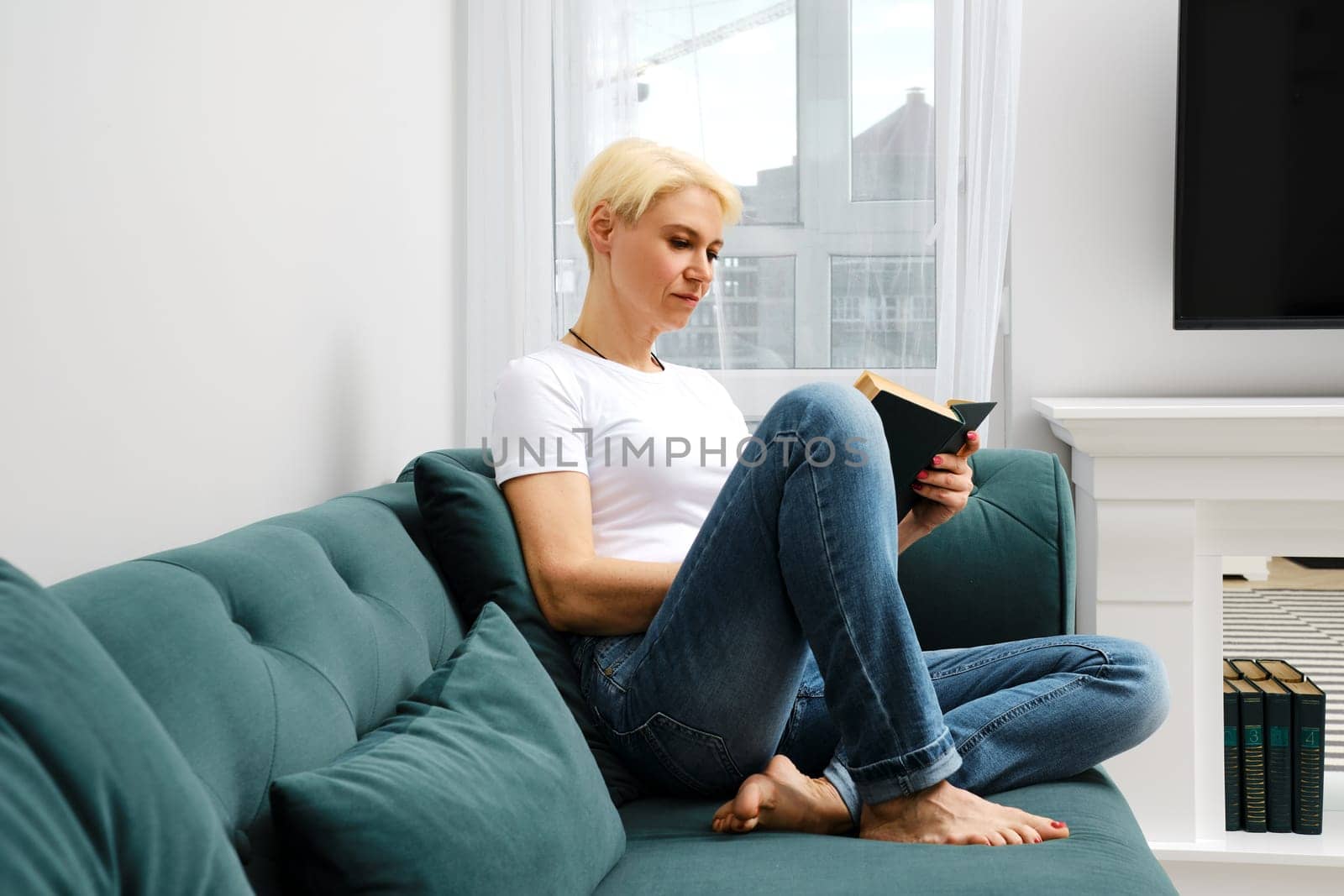 A relaxed middle-aged woman in casual clothing sits on a sofa, engrossed in her novel