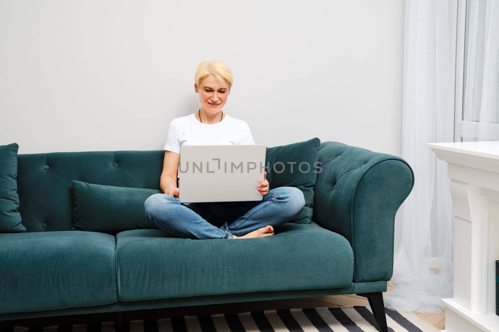 A mature woman relaxes on sofa while watching movies and browsing the web on her laptop, enjoying the comfort of independent domestic life.