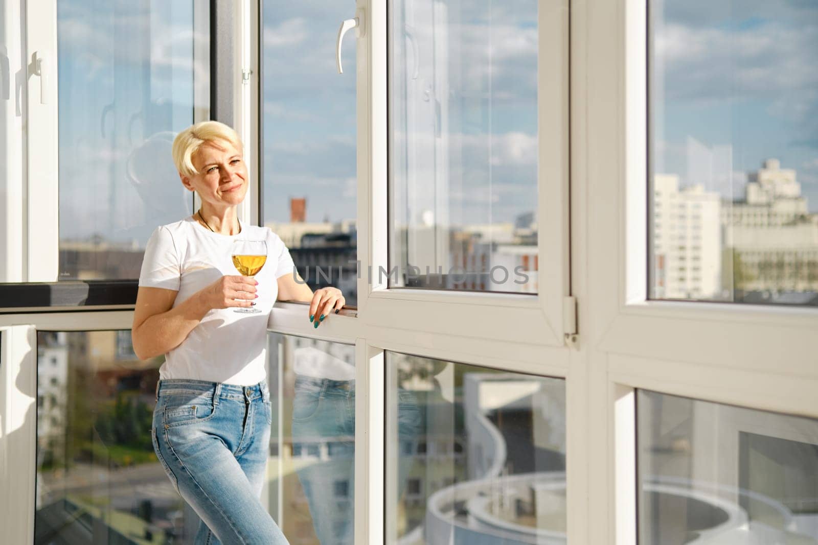 Positive middle-aged woman on balcony of high-rise apartment with glass of apple juice under bright sun