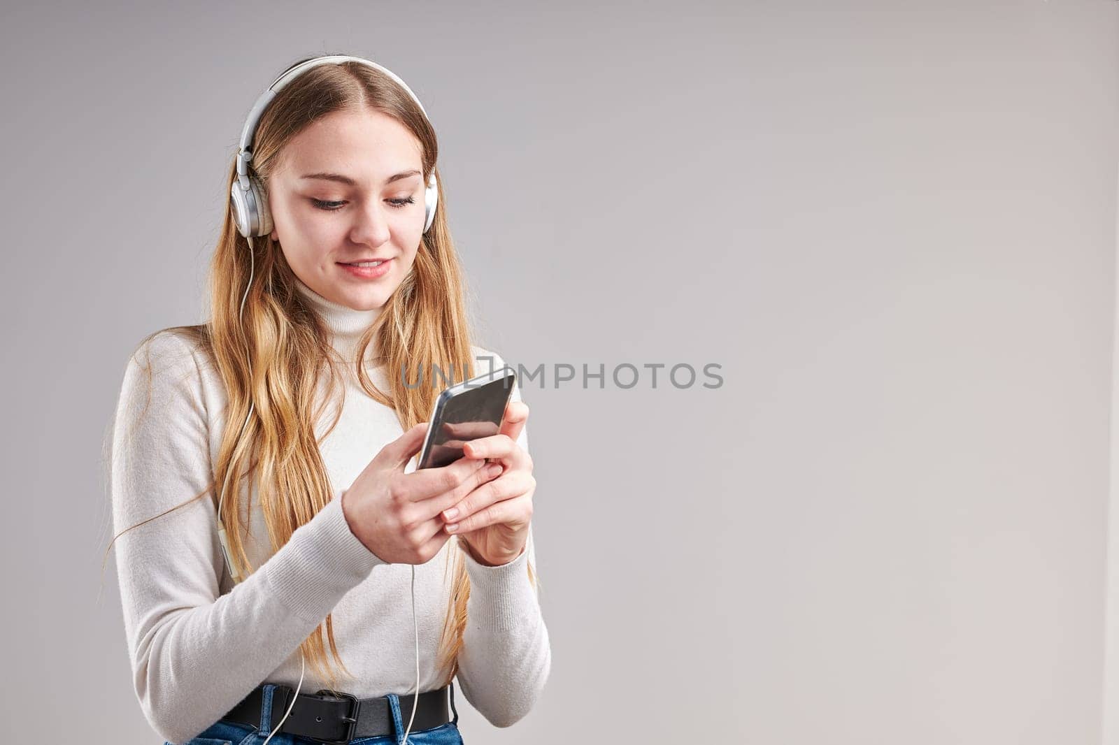 Young woman girl listening to music streaming content having fun watching video enjoying video chat talking with friends making gestures faces using smartphone earphones headphones standing over plain grey background