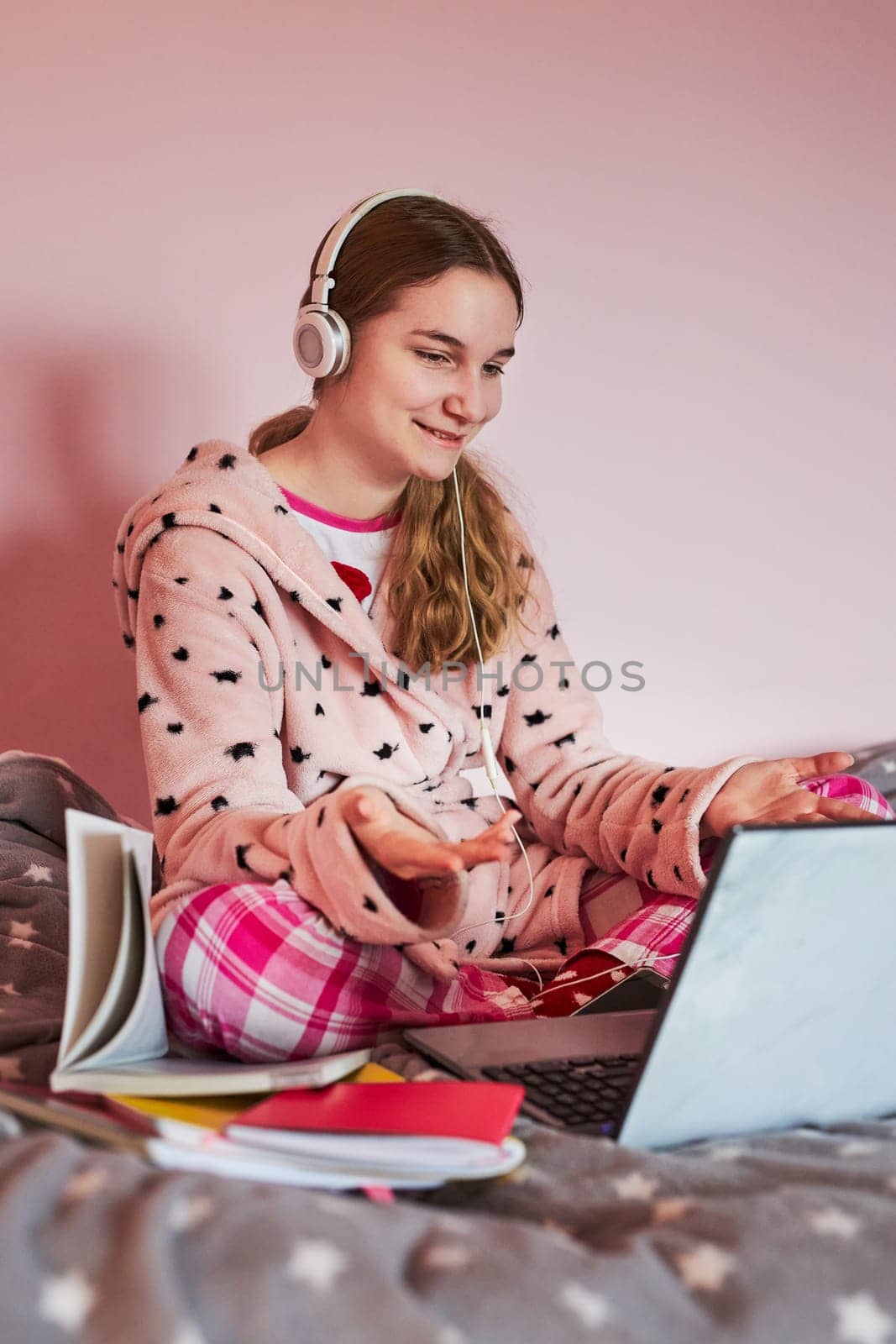 Student learning online, watching lesson remotely, listening to professor, talking with classmates on video call from home during quarantine. Young girl using laptop, headphones, books, manuals sitting in bed