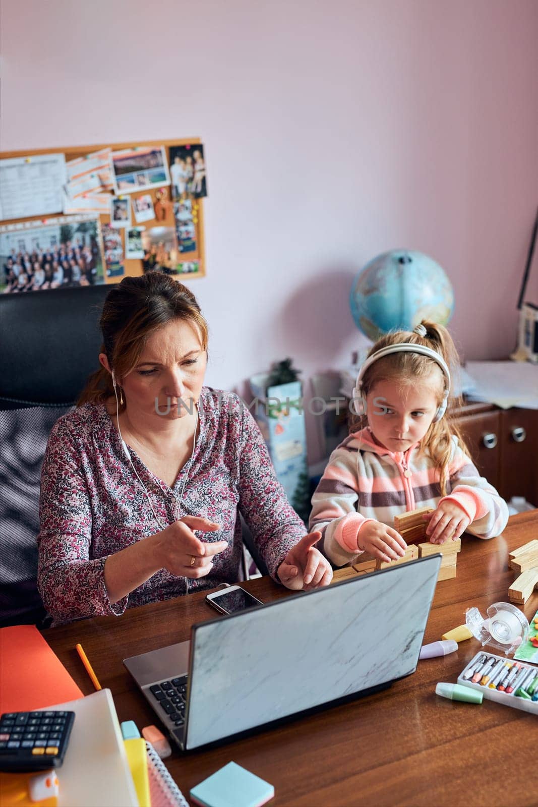 Woman mother working doing her job remotely during video chat call stream online course webinar on laptop from home while her daughter playing with bricks toy. Woman sitting at desk in front of computer looking at screen