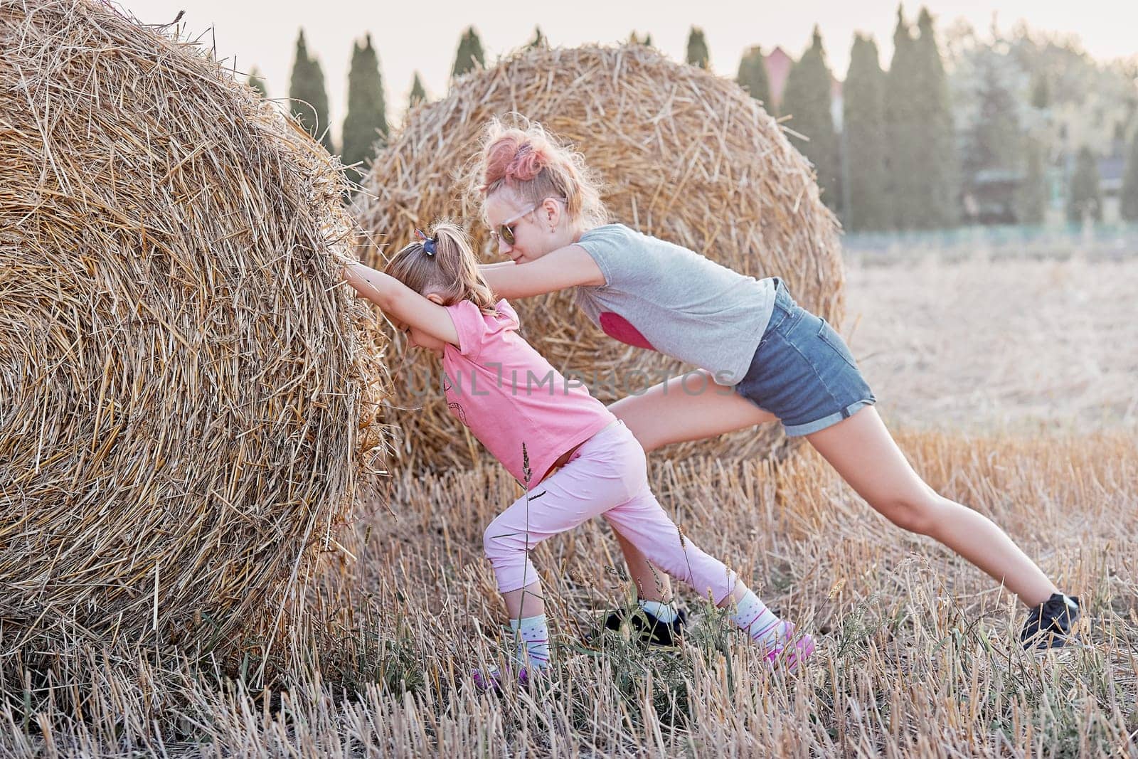 Sisters, teenage girl and her younger sister pushing hay bale playing together outdoors in the countryside. Candid people, real moments, authentic situations