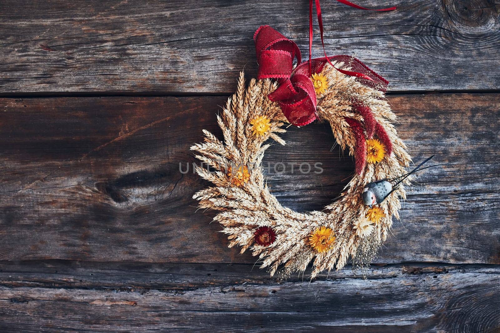 Wreath of golden ears of wheat, dried flowers and herbs tied with red ribbon hung on wooden wall. Natural rustic decoration