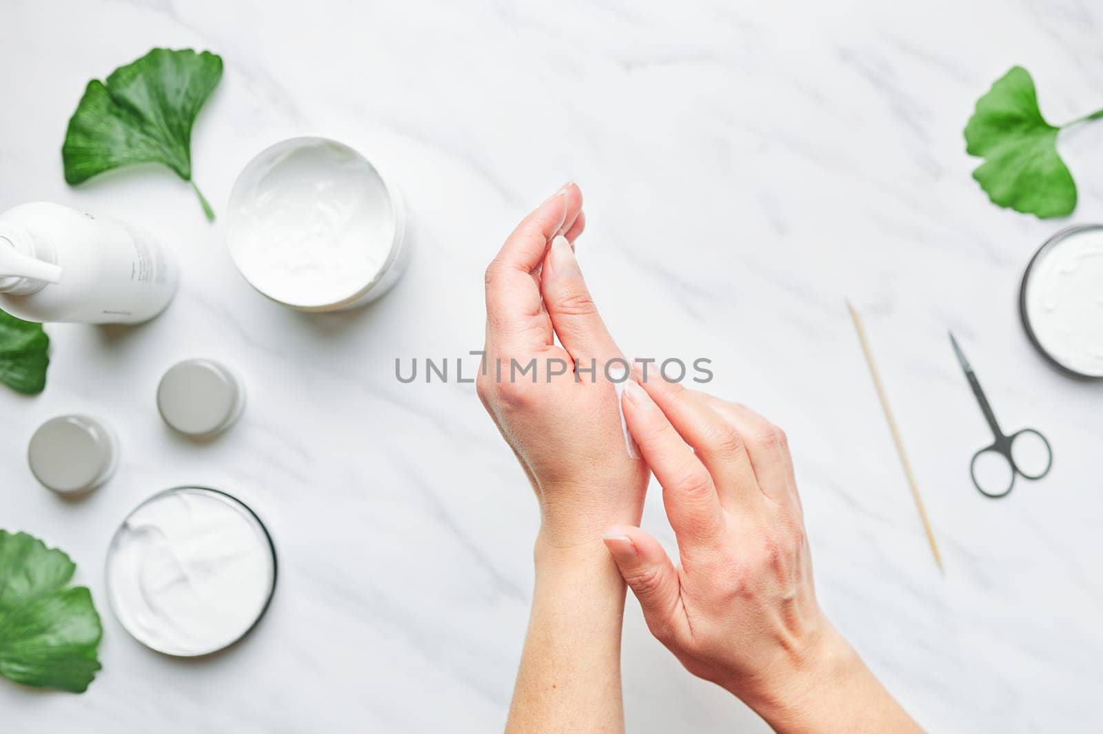 Woman applying  cosmetic moisturizing hand cream. Cosmetic products, green leaves on white table. Spa, manicure, skin care concept. Flat lay, overhead view