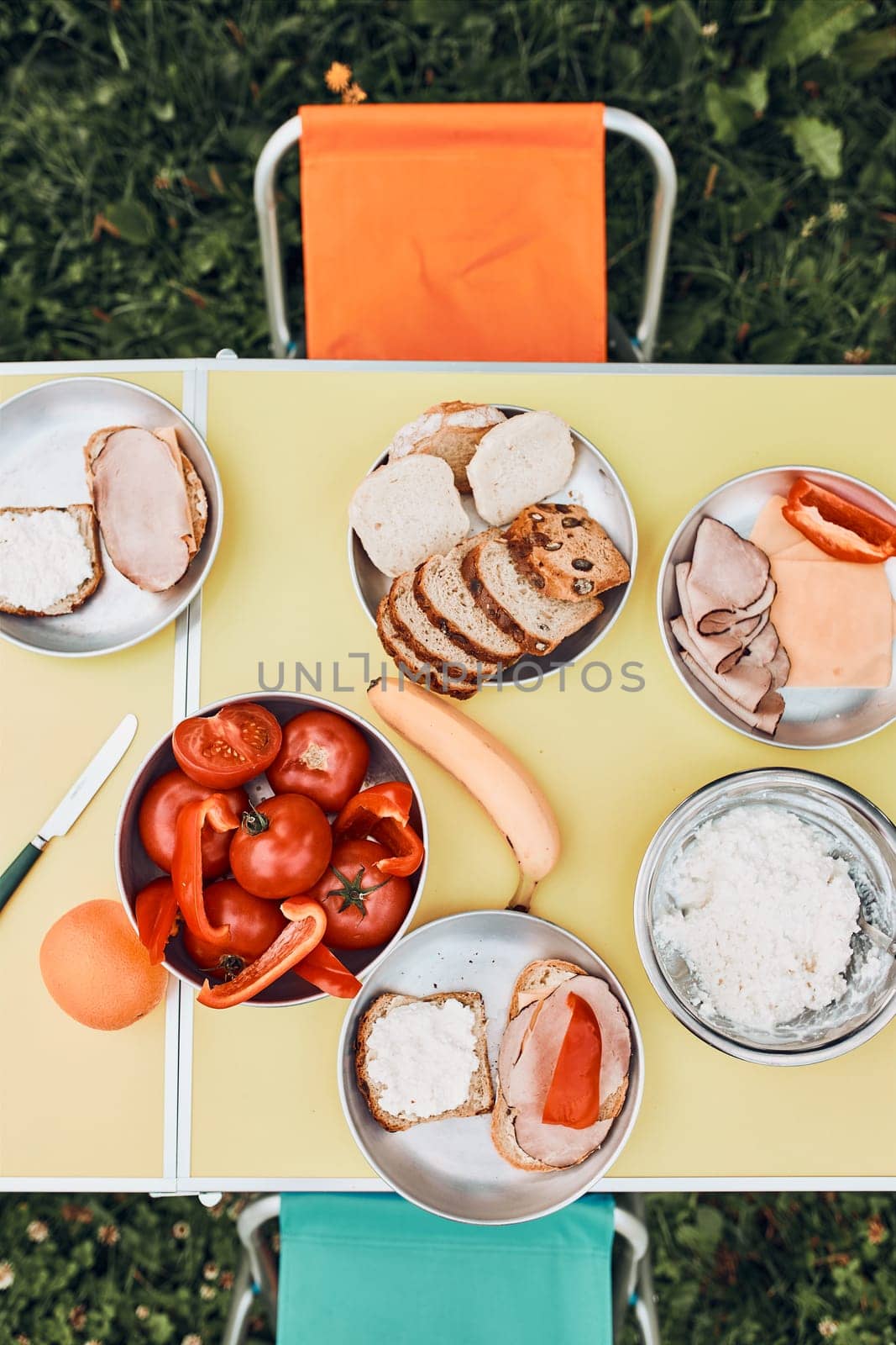 Breakfast prepared during summer vacation on camping. Bread, cottage cheese, cold meat, tomatoes, fruits and coffee cups on table. Close up of outdoor table setting from above