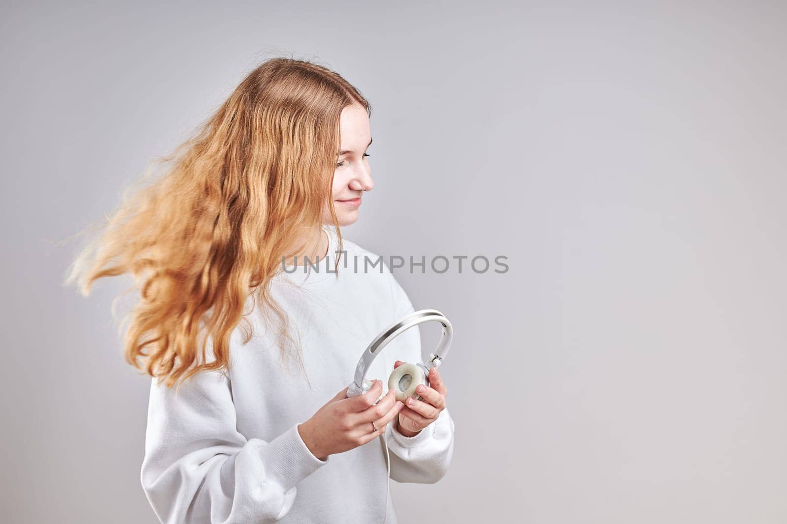 Young woman girl listening to music streaming content having fun watching video enjoying video chat talking with friends making gestures faces using smartphone earphones headphones standing over plain grey background