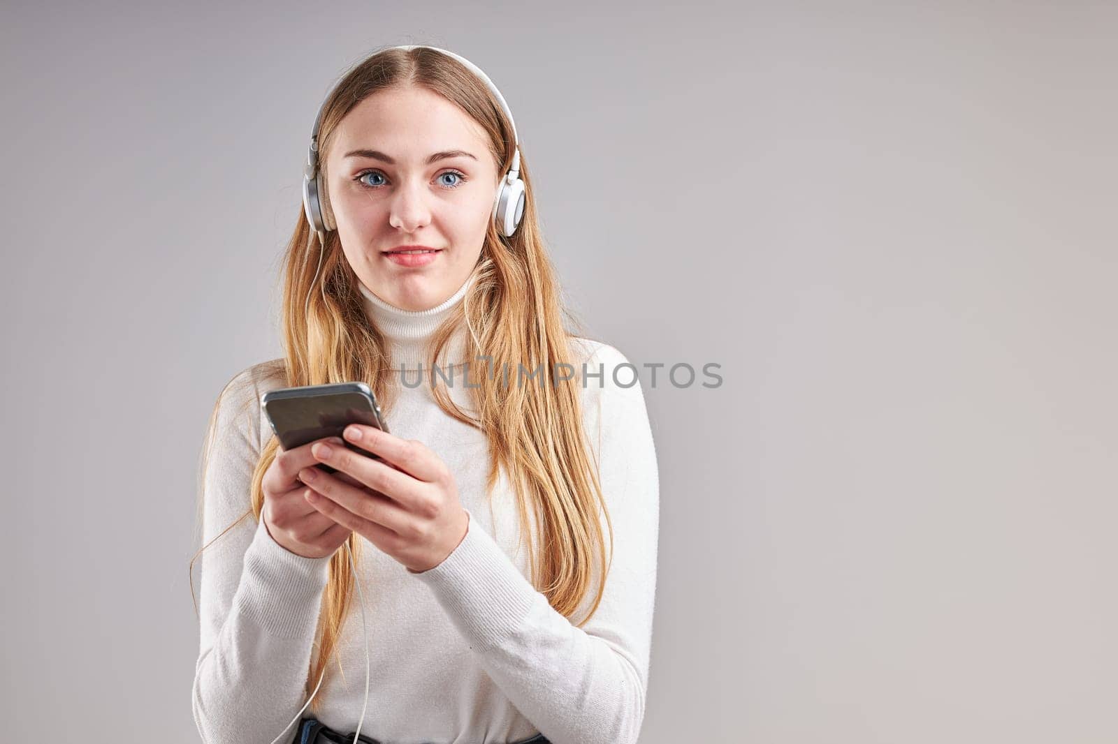 Young woman girl listening to music streaming content having fun watching video enjoying video chat talking with friends making gestures faces using smartphone earphones headphones standing over plain grey background