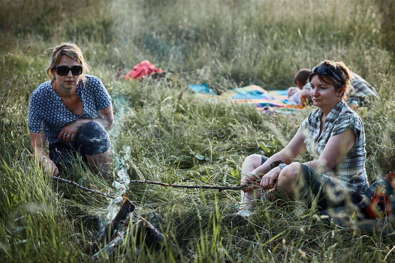 Women roasting a marshmallows over a campfire on meadow. Families spending vacations close to nature. Candid people, real moments, authentic situations