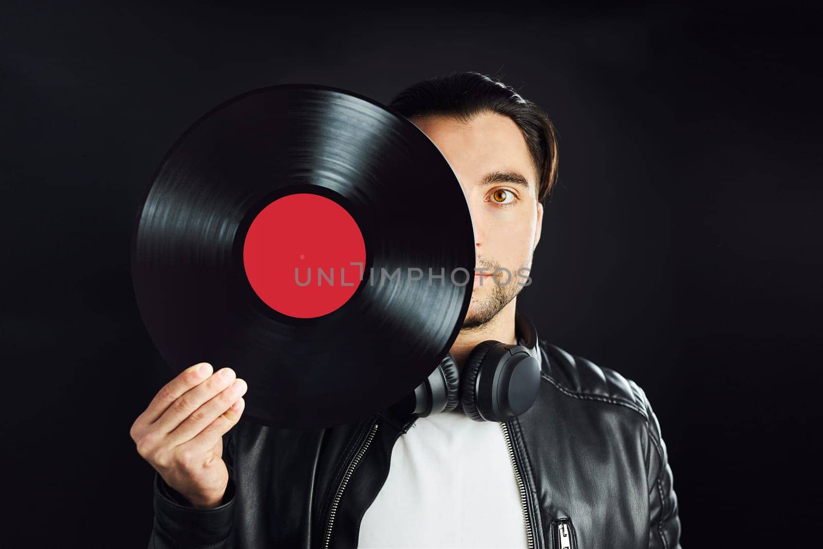 Man holding retro vinyl record covering face. Rock style. Vintage music style. Male wearing black jacket holding vinyl record disc standing on dark background. Retro music. Classic audio