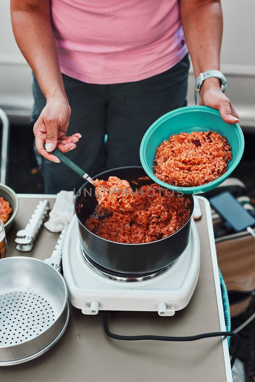 Close up of female hands putting rice dish with tomato sauce to a bowl. Woman cooking meal on electric stove on camping during summer vacations. Camping life, preparing a meal