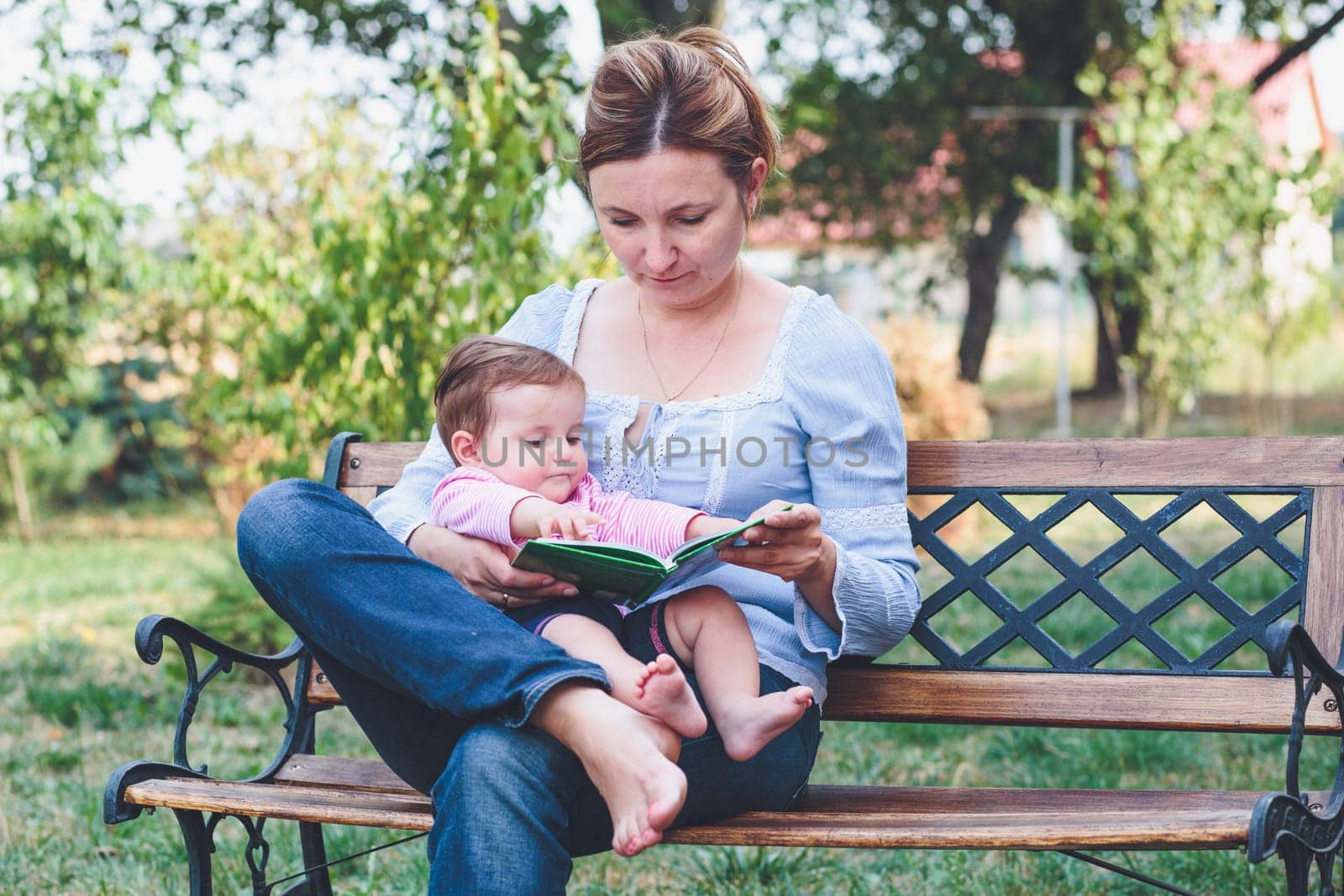 Mom reading a book and showing the pictures her little daughter sitting on bench in the garden