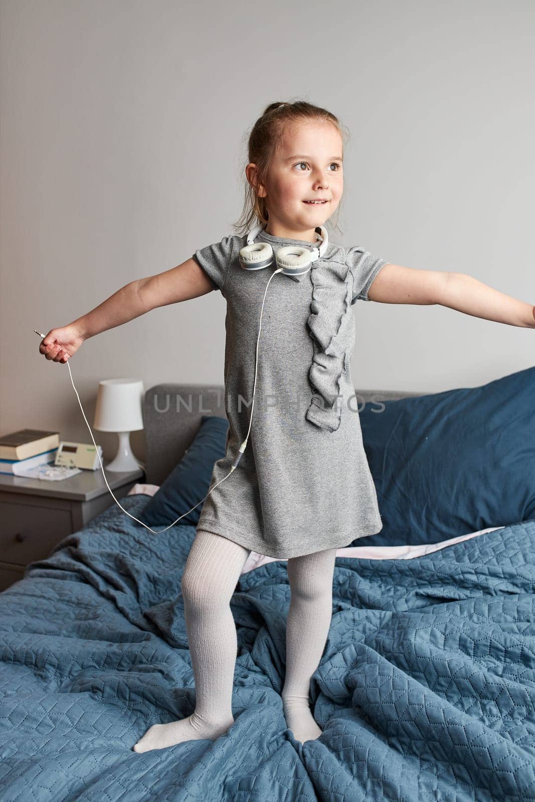 Little girl singing holding headphones cord imitating herself a real singer. Child having fun jumping dancing listening to music on bed in bedroom at home