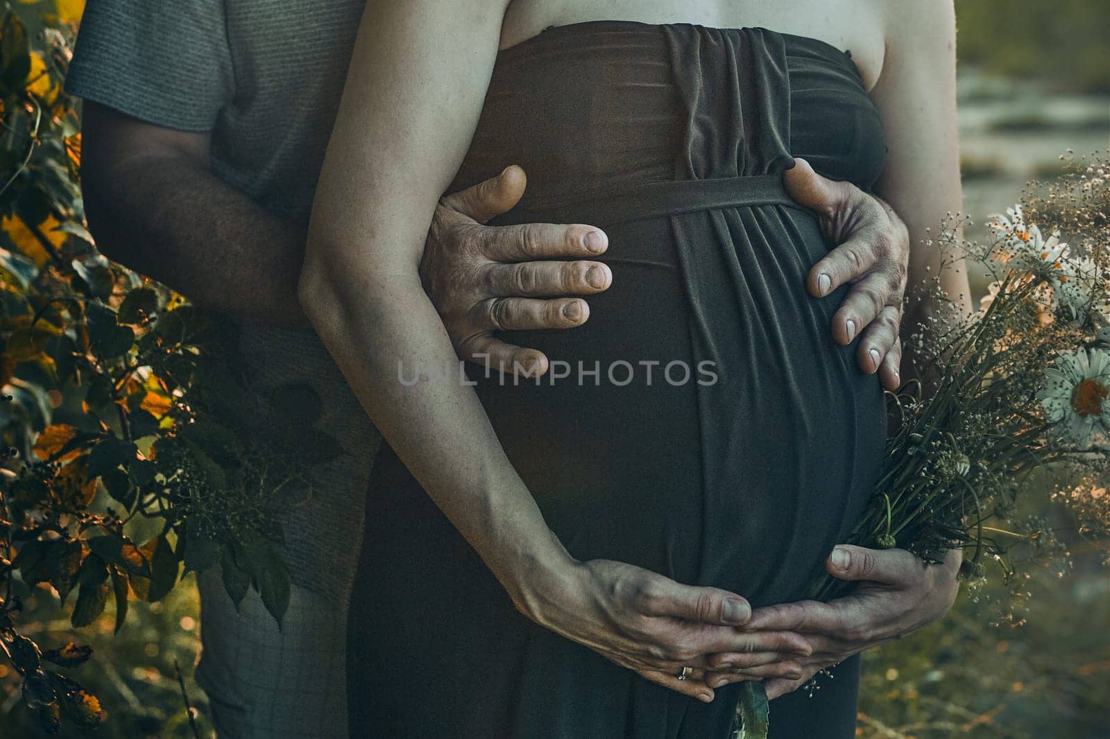 Pregnant woman and her husband hugging her tummy standing outdoors surrounded by nature. Pregnancy, expectation, motherhood concept