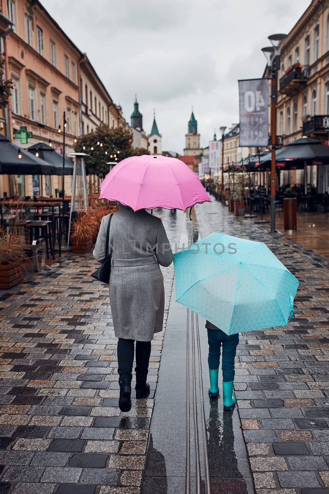 Back view of mother and her daughter holding the pink and blue umbrellas walking in a downtown on rainy gloomy autumn day