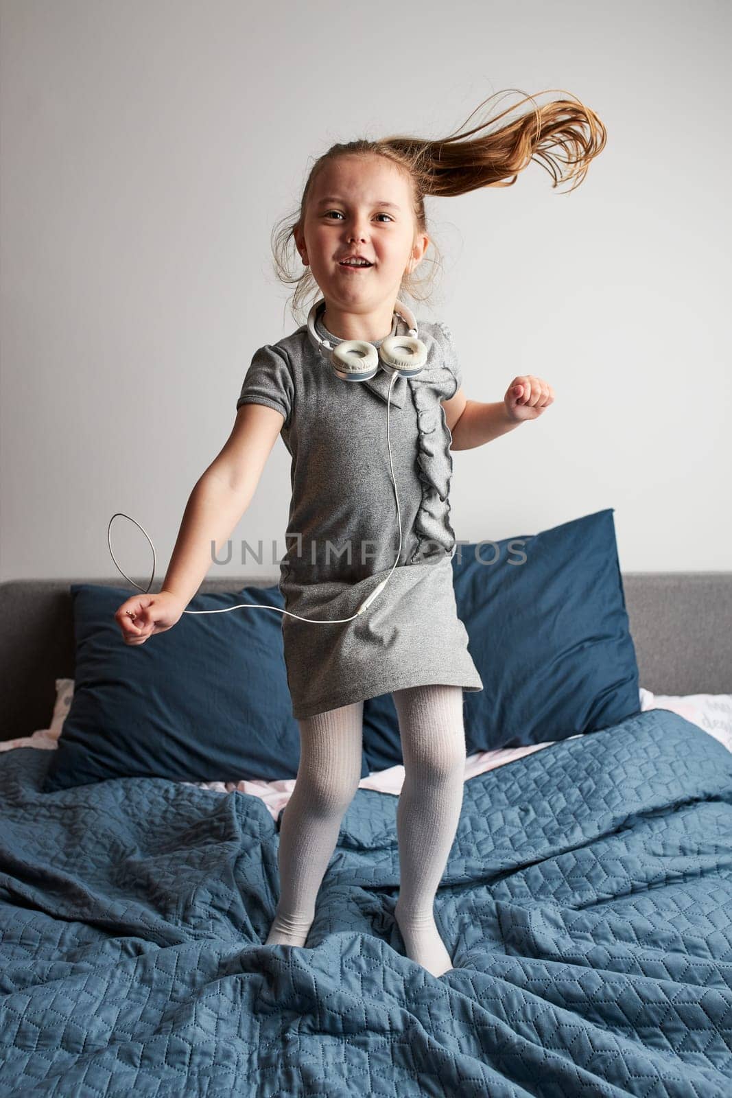 Little girl singing holding headphones cord imitating herself a real singer. Child having fun jumping dancing listening to music on bed in bedroom at home