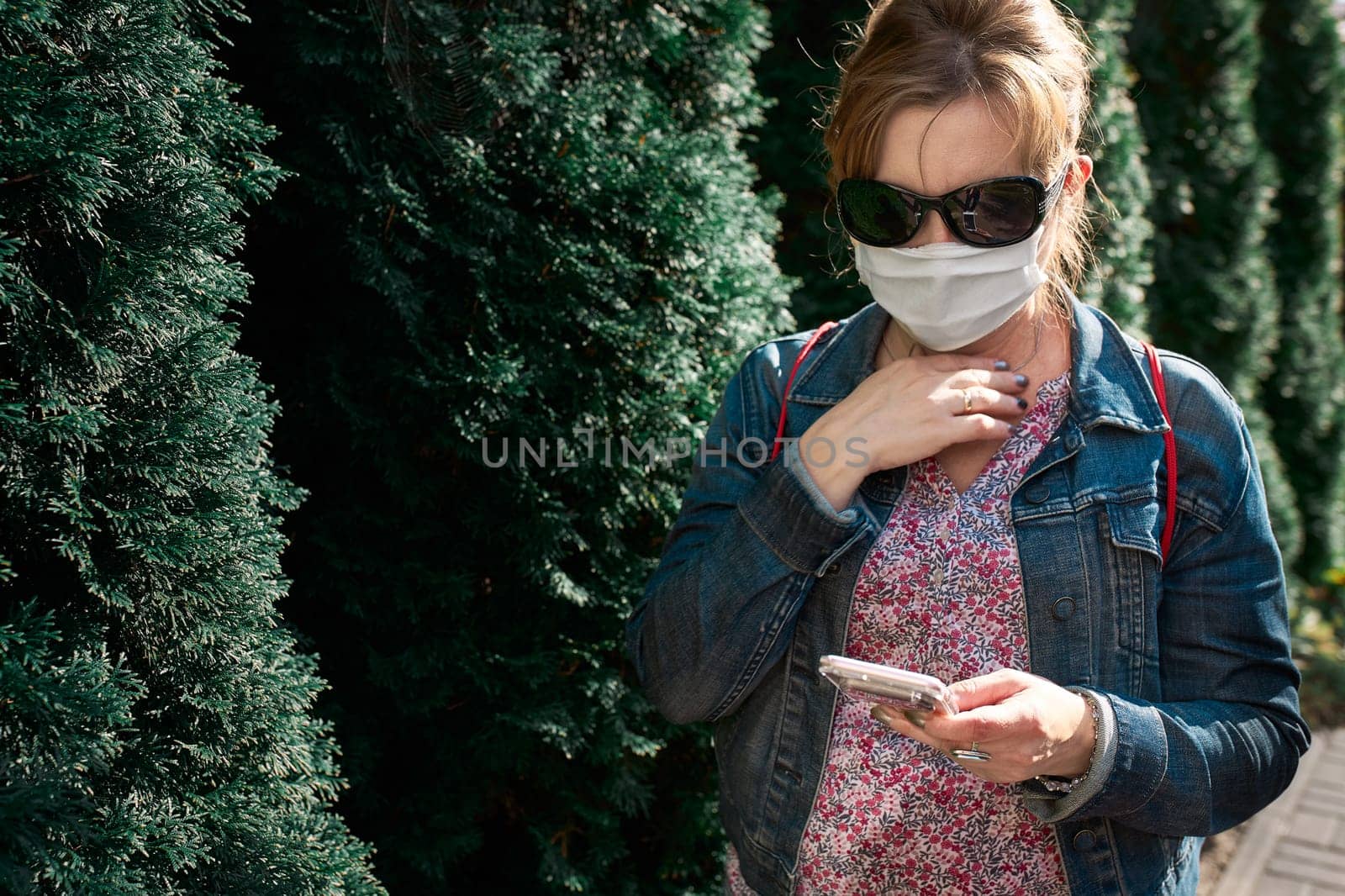 Woman using smartphone outdoors wearing face mask during pandemic coronavirus outbreak standing at front of green tress