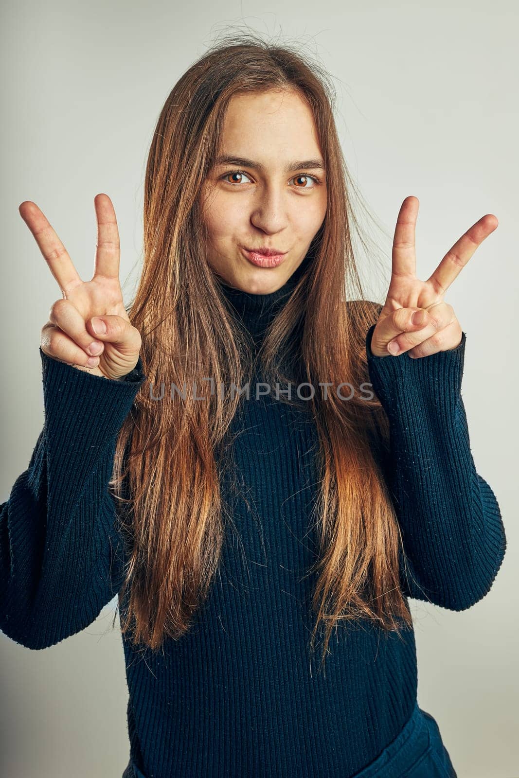 Excited girl gesturing peace sign. Portrait in positive mood. Girl making victory sign gesture. Beautiful model posing in studio over plain background looking at camera