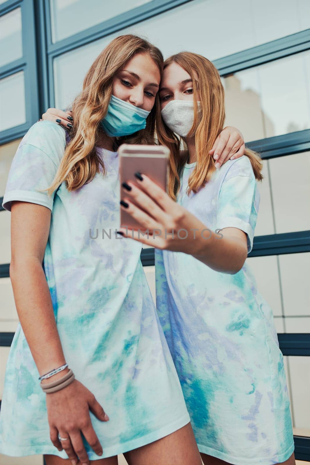 Young women girls taking selfie photos using smartphone standing at store front downtown. Girls wearing face masks to avoid virus infection and to prevent the spread of disease in time of coronavirus