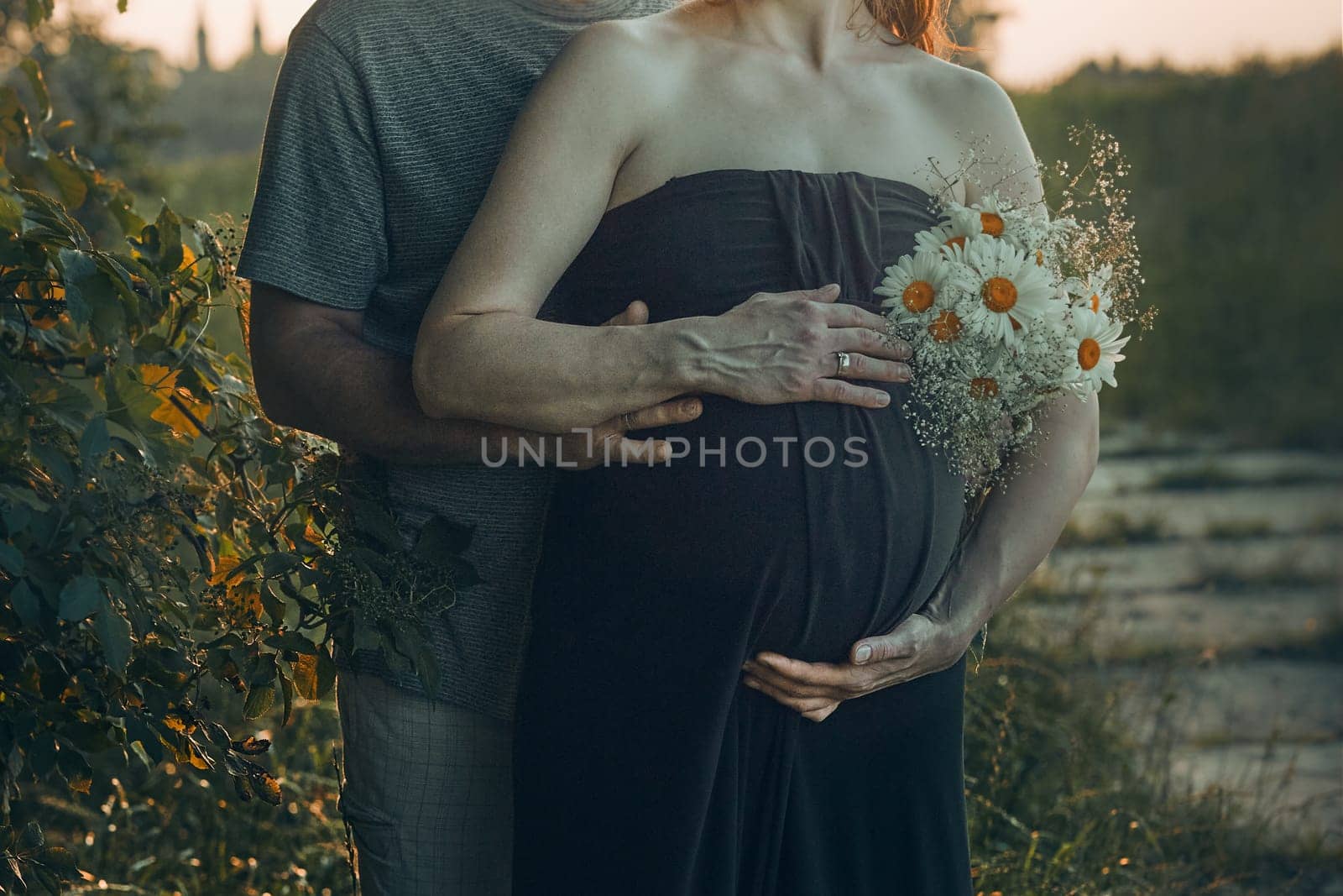 Pregnant woman and her husband hugging her tummy standing outdoors surrounded by nature. Pregnancy, expectation, motherhood concept
