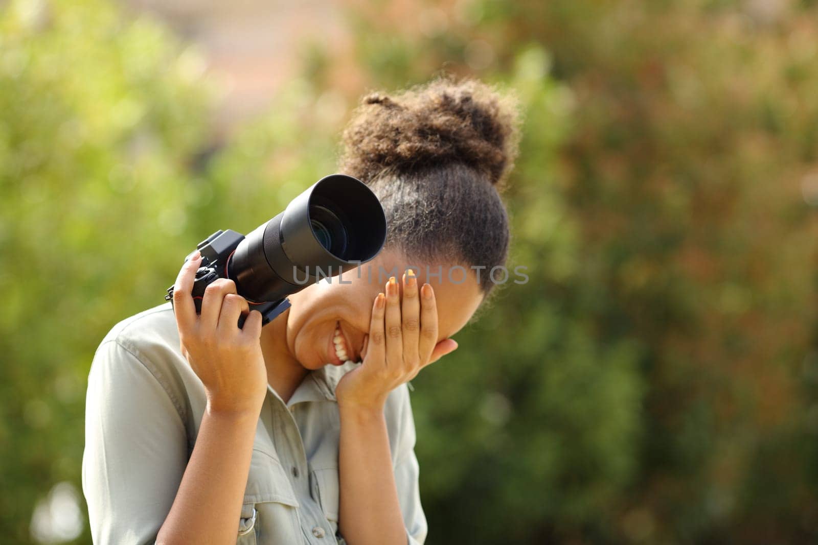 Embarrassed black photographer laughing and hiding face holding mirrorless camera in a park