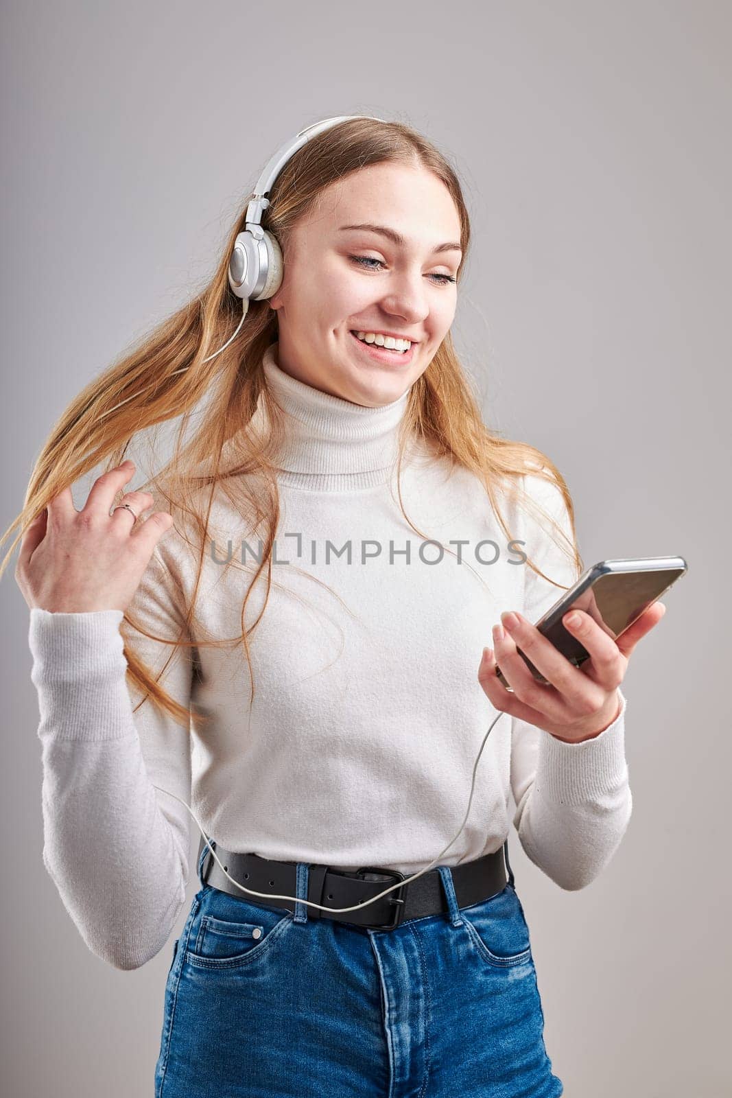 Young woman girl listening to music streaming content having fun watching video enjoying video chat talking with friends making gestures faces using smartphone earphones headphones standing over plain grey background