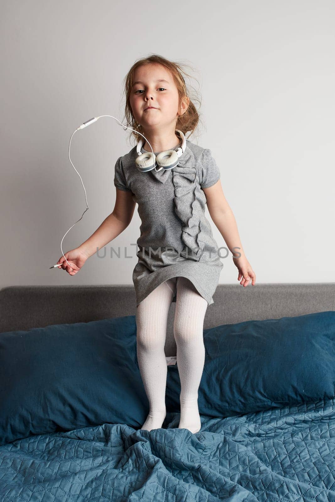 Little girl singing holding headphones cord imitating herself a real singer. Child having fun jumping dancing listening to music on bed in bedroom at home