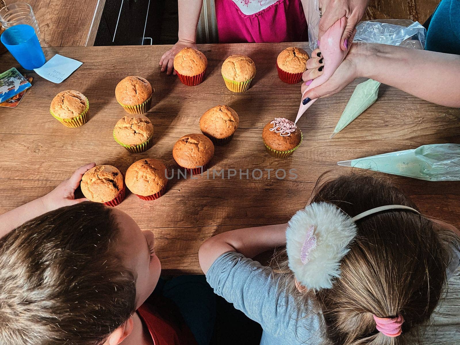 Group of children baking cupcakes, squeezing cream from confectionery bag, preparing ingredients, topping, sprinkles for decorating cookies. Kids cooking, working together in kitchen at home. Concept of happy family