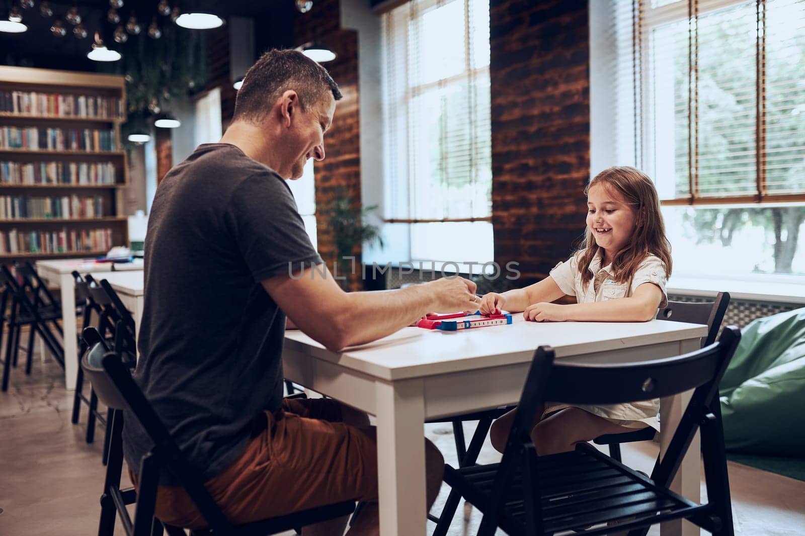Father playing board game with her daughter in public library. Child spending time with dad after classes at primary school. Positive competition. Family time