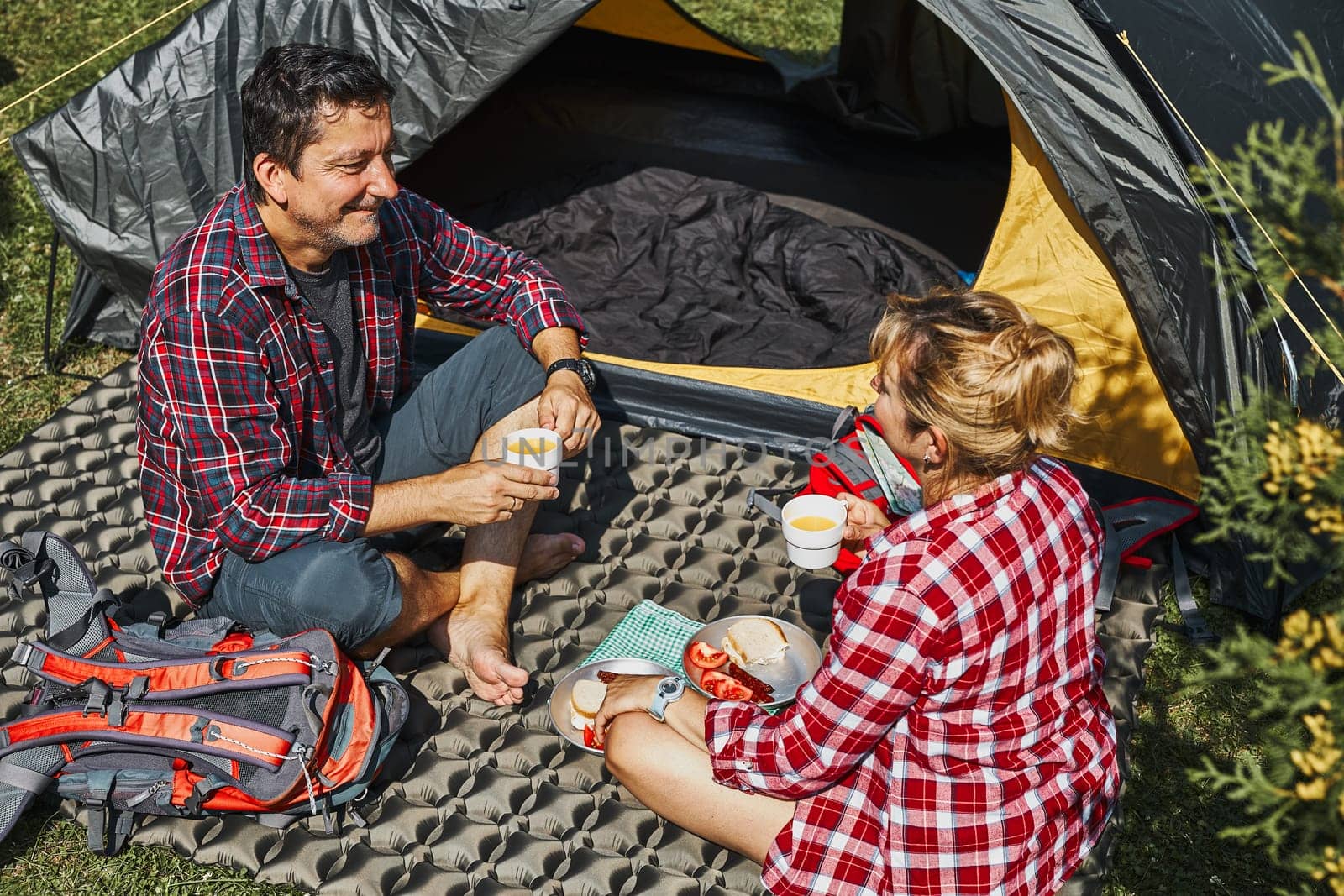 Couple having breakfast sitting by tent at camping. People actively spending summer vacations close to nature outdoors. Concept of camp life
