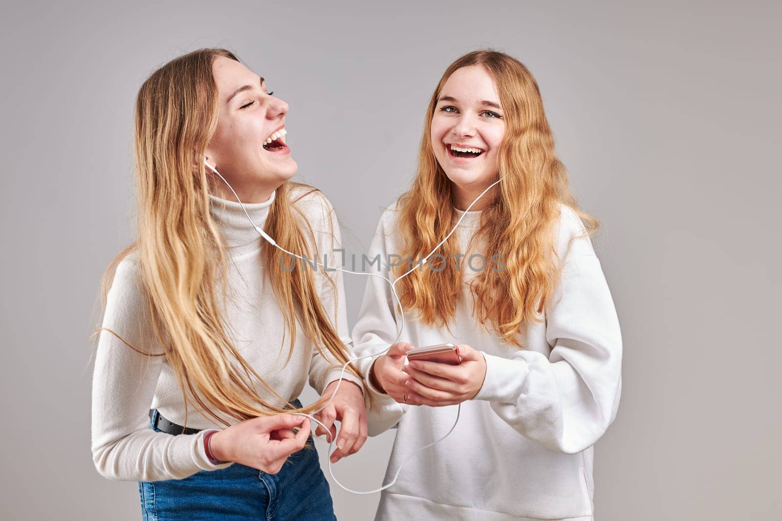 Young women girls listening to music together streaming content having fun watching video enjoying video chat talking with friends making gestures faces using smartphone earphones headphones standing over plain grey background