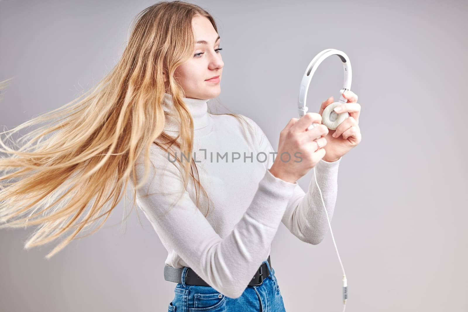 Young woman girl listening to music streaming content having fun watching video enjoying video chat talking with friends making gestures faces using smartphone earphones headphones standing over plain grey background
