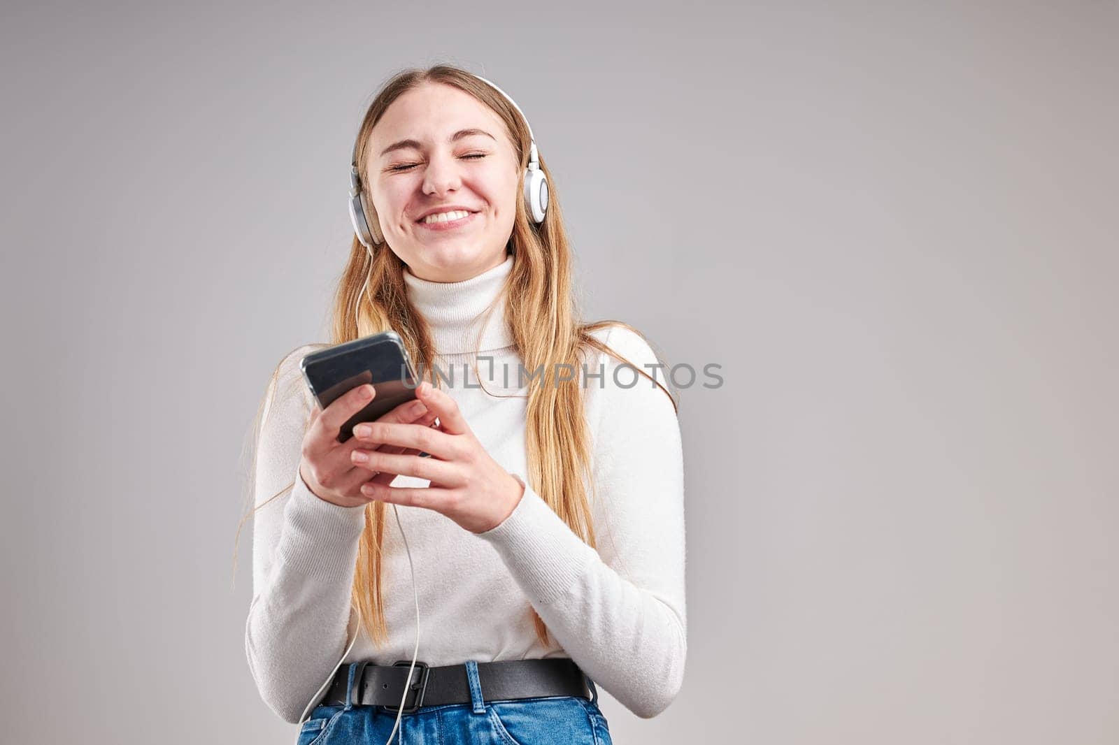Young woman girl listening to music streaming content having fun watching video enjoying video chat talking with friends making gestures faces using smartphone earphones headphones standing over plain grey background