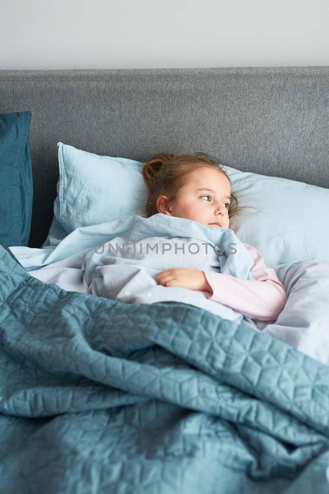 Ill little girl lying in bed covered with duvet. Sick kid recovering, resting in bed, looking at window