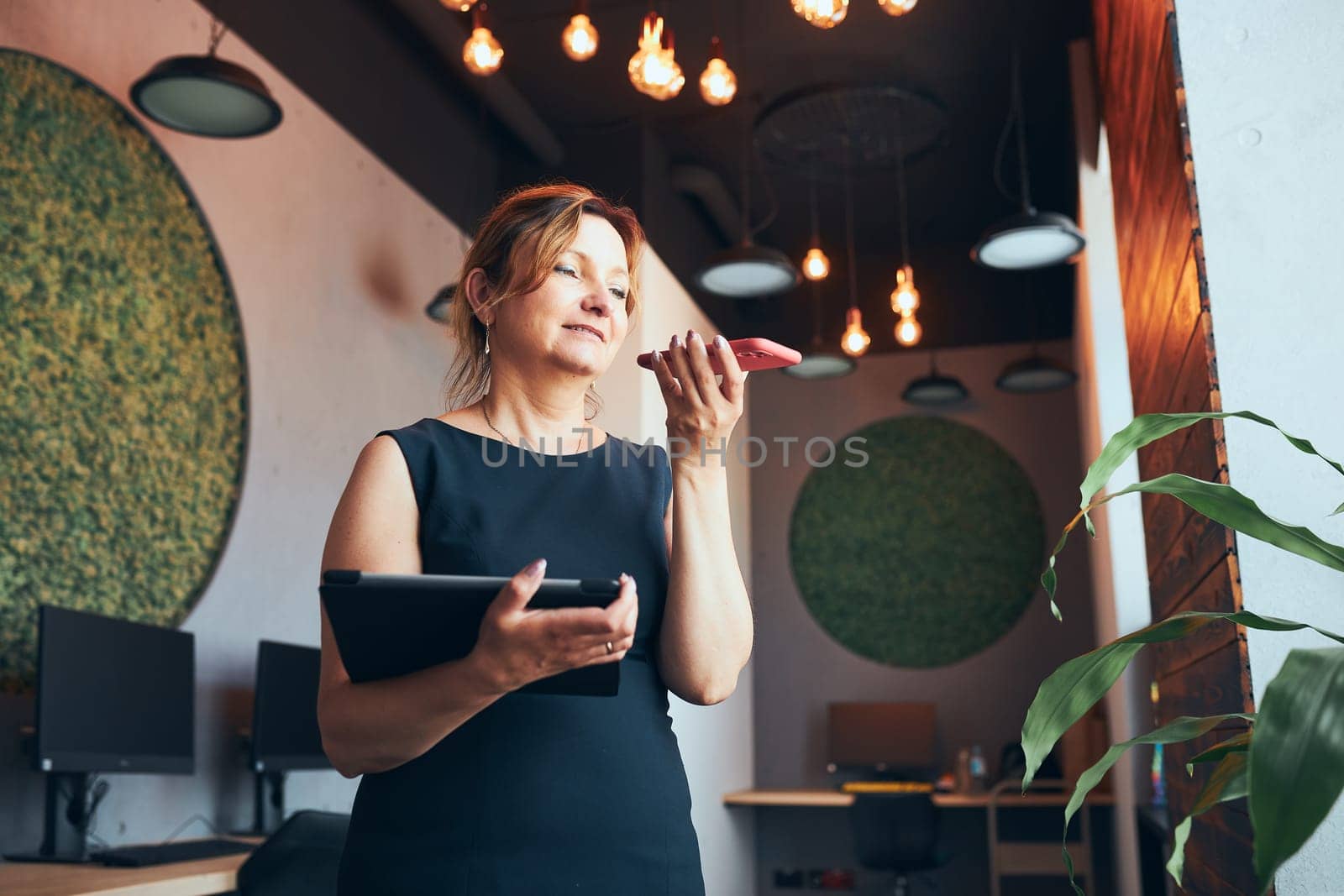 Businesswoman having business call working on tablet in office. Mature busy woman using touch pad computer standing by window in modern interior. Female manager focused on work holding digital device. Using technology