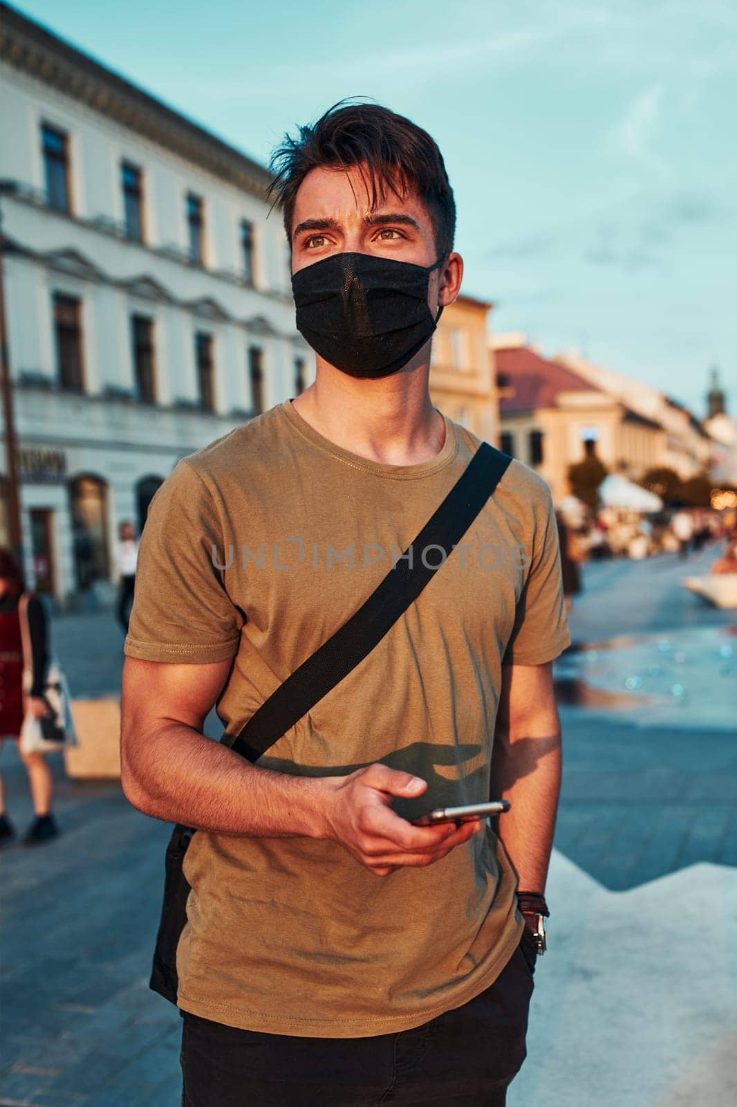 Young man standing in the city center in the evening, looking away, wearing the face mask to avoid virus infection and to prevent the spread of disease in time of coronavirus