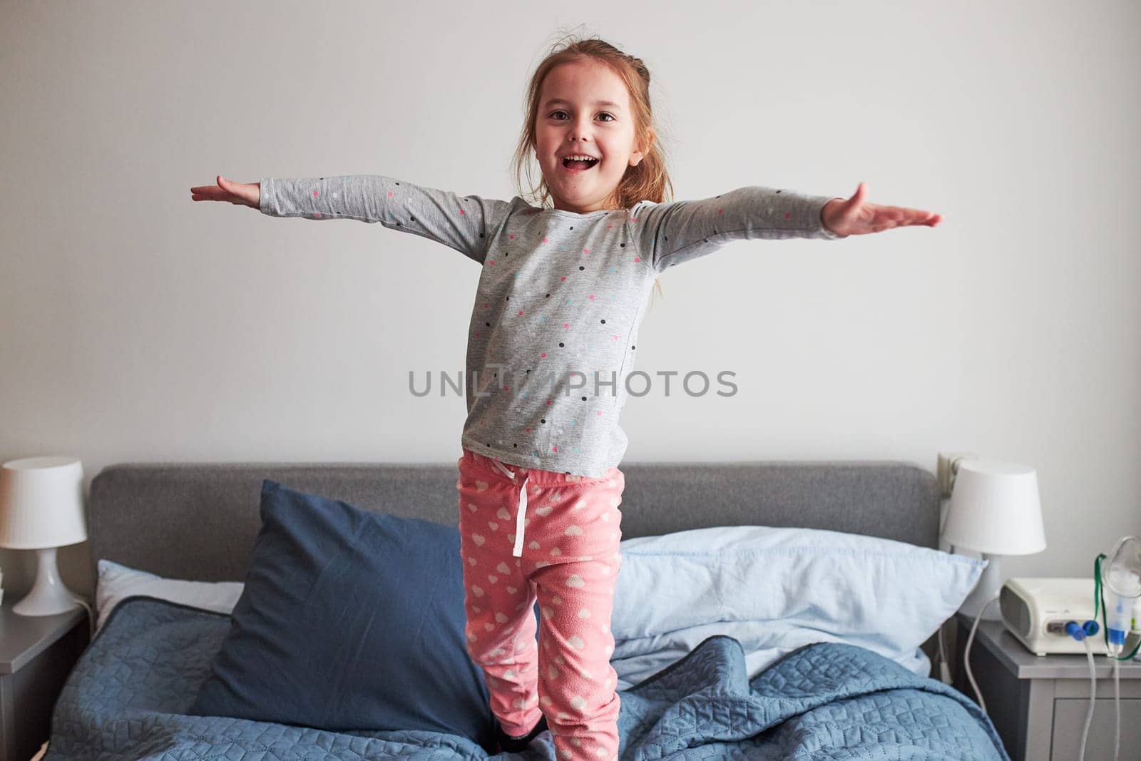 Little happy playful kid girl having fun making silly funny faces jumping on bed in bedroom in the morning at home