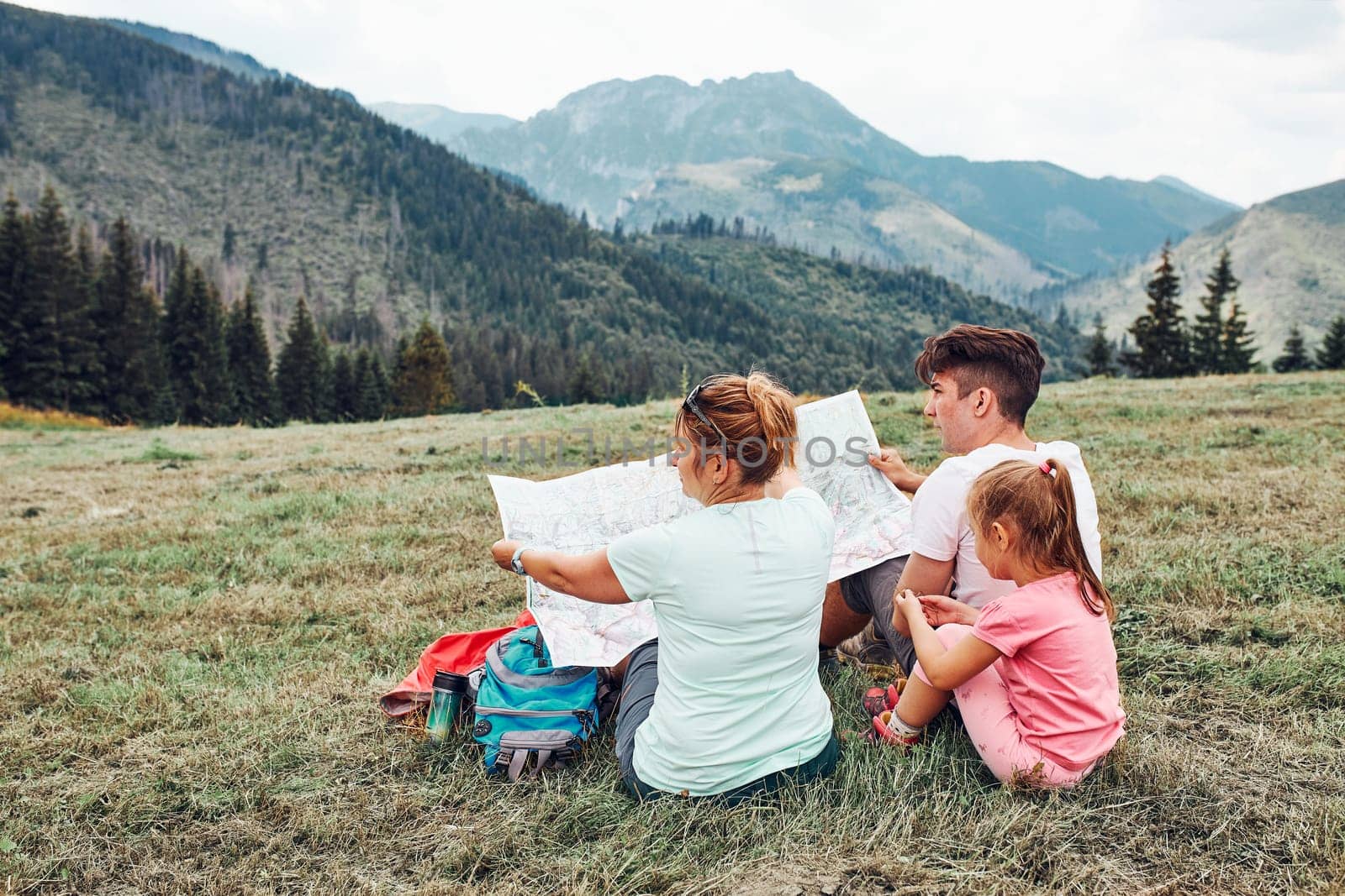 Family examining a map of mountains trials sitting on grass enjoying summer day during vacation trip in mountains. People actively spending time outside. Mountain landscape in the background