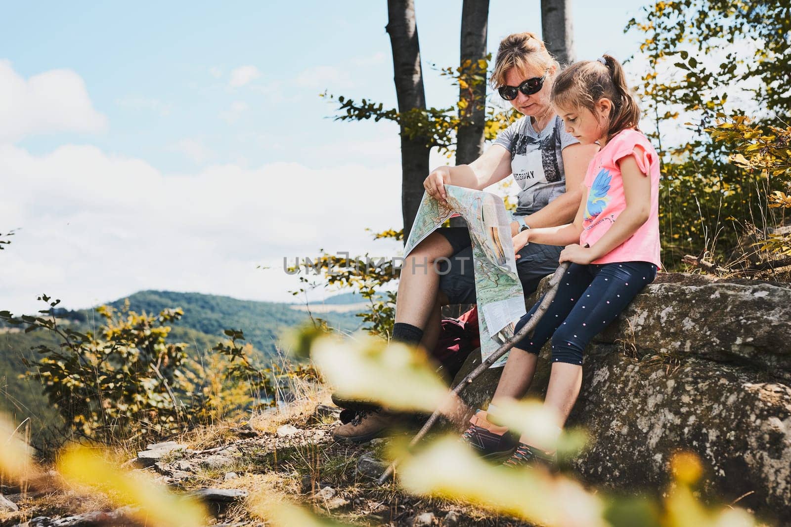 Family trip in mountains. Mother and her daughter examining a map of mountains trials sitting on rock enjoying summer day during vacation trip in mountains. People actively spending time outside