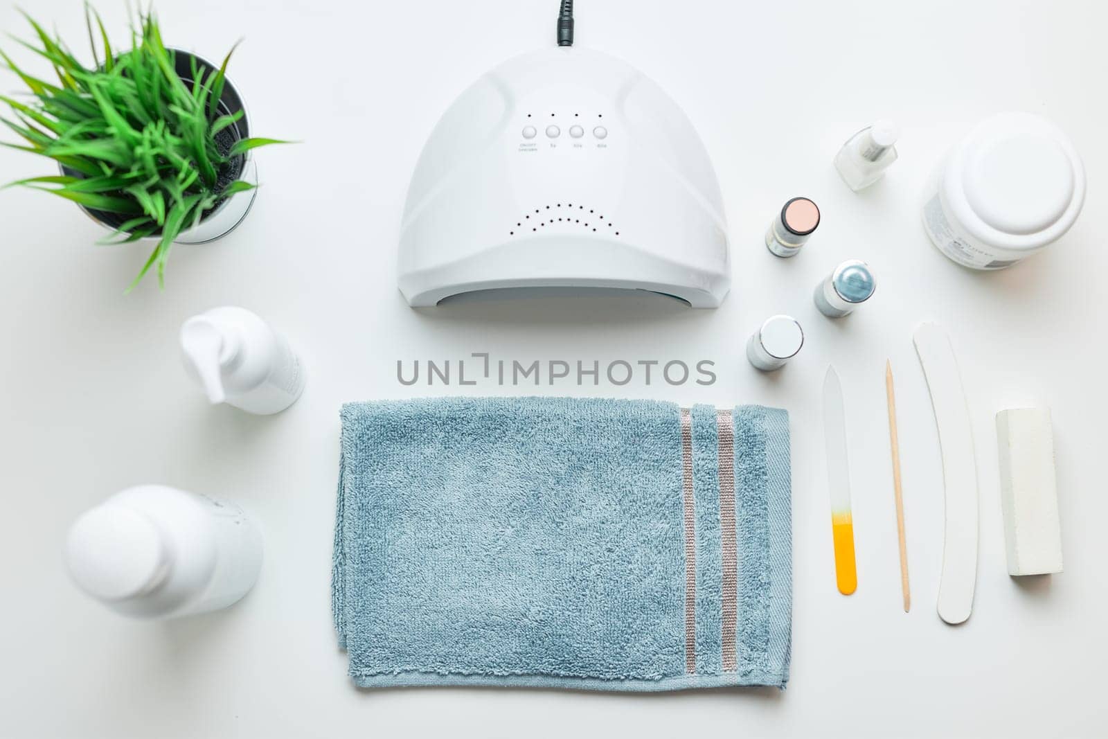 Cosmetic products set, nail hybrid gels, UV lamp, green leaves, blue towel on white table. Spa, manicure, skin care concept. Flat lay, overhead view