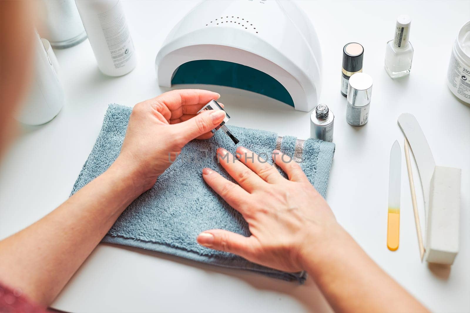 Woman applying gel hybrid polish using UV lamp. Beauty wellness spa treatment concept. Cosmetic products, UV lamp, green leaves on white table. Spa, manicure, skin care concept. Flat lay, overhead view