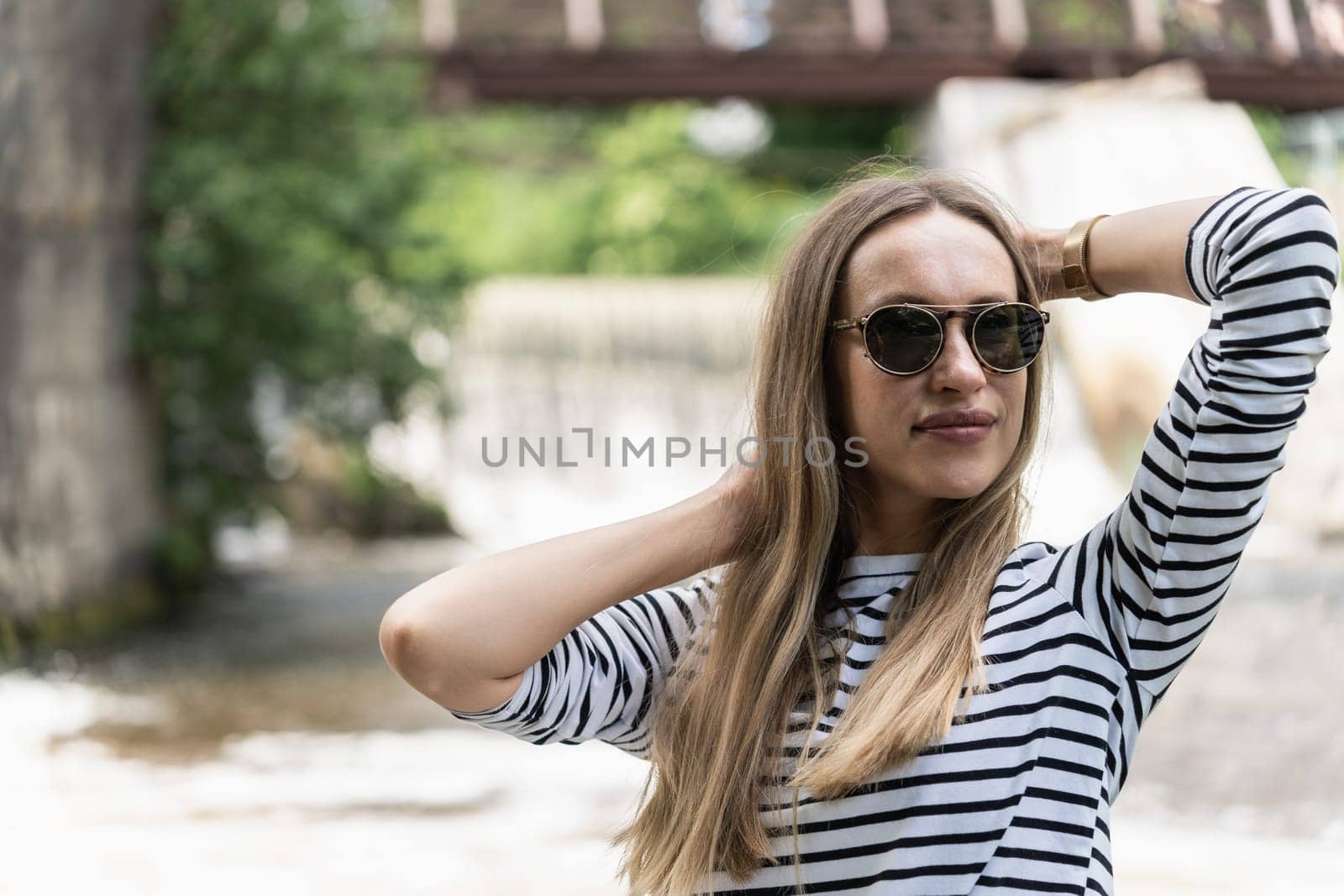 A woman in sunglasses and a striped top holds her hair near the river with her hands.