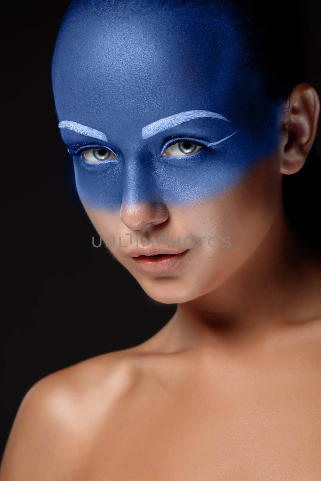 Portrait of a young woman who is posing covered with blue paint in the studio on a black background.