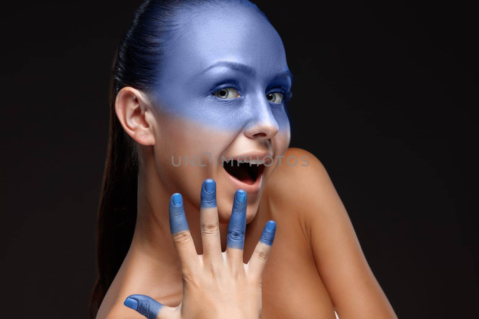 Portrait of a smiling young woman who is posing covered with blue paint in the studio on a black background. girl&#39 s hand around the face and painted blue