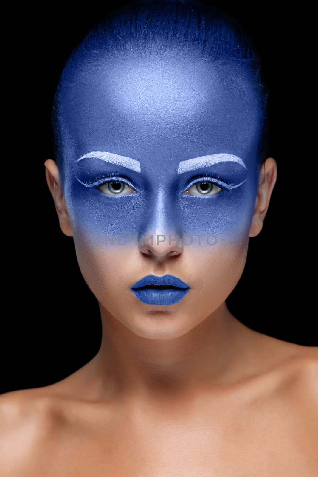 Portrait of a young woman who is posing covered with blue paint in the studio on a black background. Girl has blue lips and white eyebrows